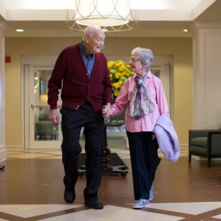 An elderly couple holding hands and walking together indoors.