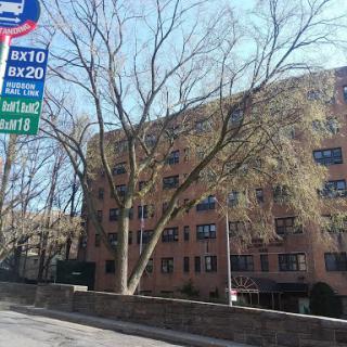 A bus stop with route information in an urban setting, featuring a tree and a nearby building.