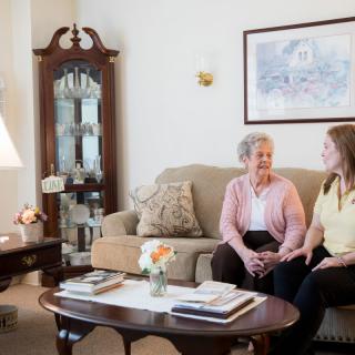 An elderly woman and a caregiver sitting on a couch in a cozy living room, engaged in conversation.