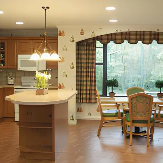 A traditional kitchen with wood cabinetry and an island counter, featuring a dining area with floral wallpaper and checked curtains.