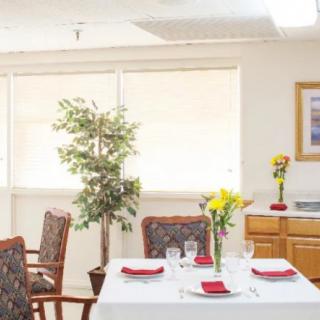 A bright dining room with a neatly set table surrounded by chairs, featuring floral decorations and a large window.