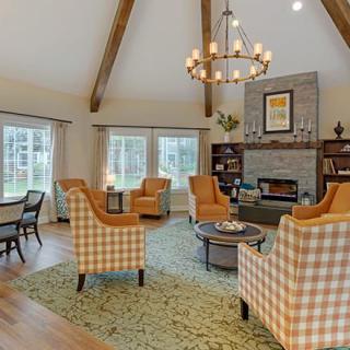 A cozy living room with a high ceiling featuring wood beams, comfortable armchairs, a fireplace, and large windows letting in natural light.