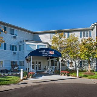 The image shows a three-story building with an entrance featuring a blue awning. Trees are visible along the pathway to the entrance.