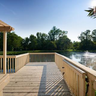 A wooden deck overlooking a serene lake with a fountain and surrounding trees in bright sunlight.