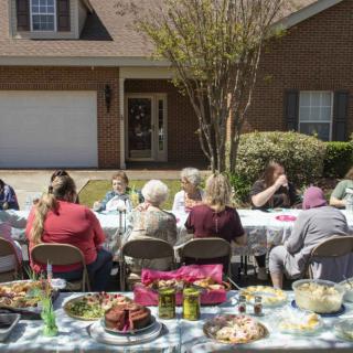 A family gathering around a table with various dishes in front of a house on a sunny day.
