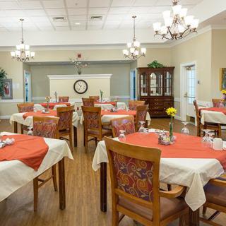 A cozy dining room with multiple tables set with white and orange tablecloths, surrounded by wooden chairs, illuminated by chandeliers.