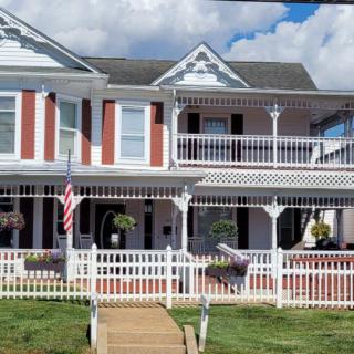 A two-story house with a porch and balcony, featuring a white picket fence and an American flag under a cloudy sky.