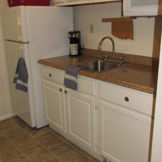 A small kitchen area featuring a refrigerator, sink, countertop, and cabinets with a coffee maker and towels.