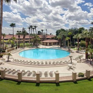 A luxurious resort featuring a large circular swimming pool surrounded by palm trees and lounge chairs under a partly cloudy sky.