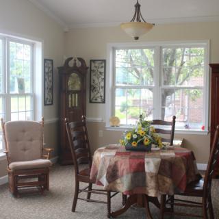 A cozy dining room with a table, chairs, and a grandfather clock, illuminated by natural light from large windows.