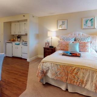 A cozy bedroom with a neatly made bed, decorative pillows, and framed artwork on the wall. The room features a side table with a lamp and a small kitchenette area.