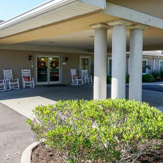 The image shows the entrance of a building with a covered porch, columns, and rocking chairs. A shrub is visible in the foreground.