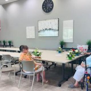 A spacious meeting room with a few seniors seated around tables, decorated with flowers and artwork, featuring a large clock on the wall.