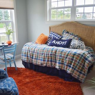 A cozy bedroom featuring a plaid-bedecked daybed with nautical-themed pillows, an orange rug, and a small table next to a chair by the window.