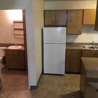 A small apartment kitchen with wooden cabinets and a white refrigerator, adjacent to a bathroom.