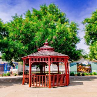 A vibrant red gazebo sits in a sunny park, surrounded by lush green trees and a clear blue sky.