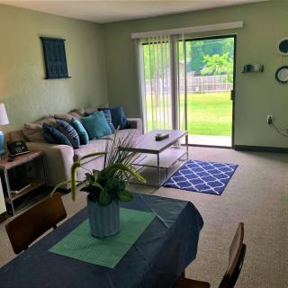 A cozy and well-lit living room with a sofa, coffee table, and dining area, opening to a green outdoor space through sliding glass doors.