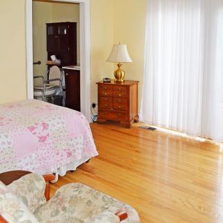 A cozy bedroom with wooden floors, featuring a bed, dresser, and a lamp by the window.