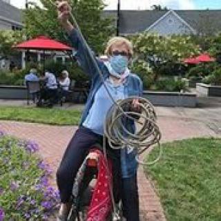 A woman wearing a mask poses playfully with a coiled rope while sitting on a bicycle in a park setting.