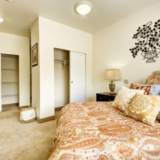 A cozy bedroom with a patterned bedspread, decorative pillows, and a lamp on the nightstand. A closet with sliding doors is visible in the corner.