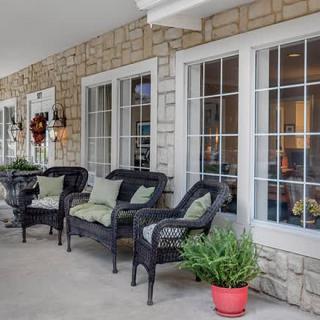A cozy porch with wicker chairs and potted plants alongside a stone wall and large windows.
