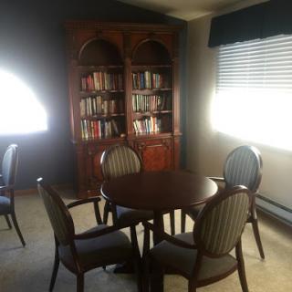 A cozy reading nook featuring a round table surrounded by upholstered chairs and a wooden bookshelf filled with books.