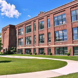 A large brick school building with numerous windows, surrounded by a green lawn and clear sky.