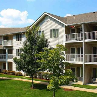 A sunny view of a three-story apartment building with balconies, surrounded by trees and a well-maintained lawn.