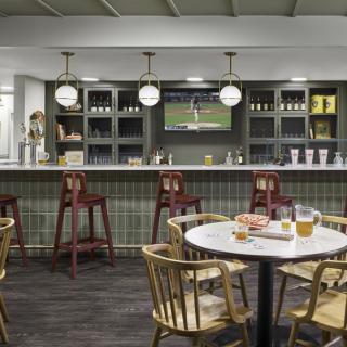 A cozy bar interior with a wooden table and chairs, featuring a well-stocked counter and a TV displaying sports.