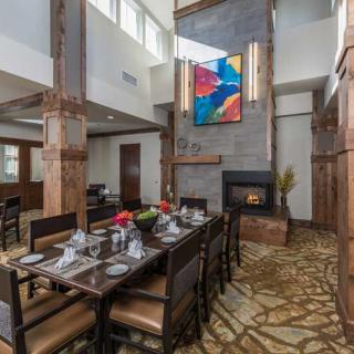 A modern dining room features a long table with elegant place settings, highlighted by large windows and an abstract painting above the fireplace.