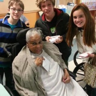 A group of children visiting an elderly person in a wheelchair in a hospital setting.