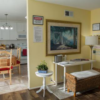 A cozy living space with yellow walls, featuring a painting, wicker furniture, and a view into the kitchen.