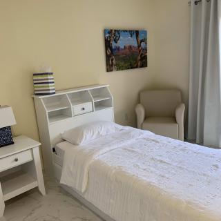 A tidy bedroom featuring a single bed with white linens, complemented by a side table and lamp, an armchair, and wall art.