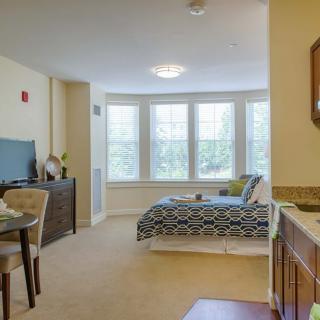 A cozy studio apartment with a bed, dining table, and wooden furniture illuminated by natural light.