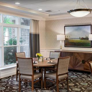 A cozy dining room with a table set for four, featuring elegant decor and ample natural light from large windows.