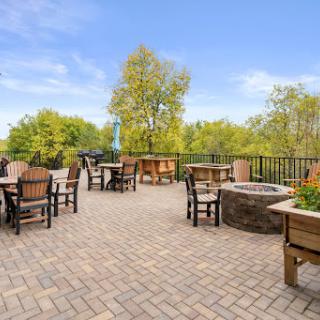 A serene outdoor patio area featuring comfortable seating and a fire pit, surrounded by lush greenery and clear blue skies.