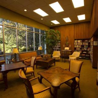 A cozy library reading room with wooden furniture and large windows letting in natural light.