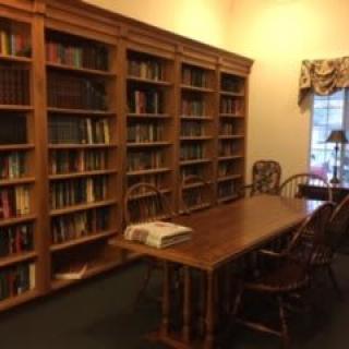 A cozy library room featuring wooden bookshelves filled with books, a large table, and several chairs for reading.