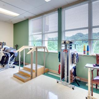 A well-equipped physical therapy room featuring exercise machines, a staircase for rehabilitation, and large windows allowing natural light.