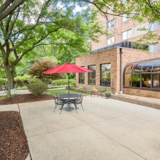 A serene outdoor patio area featuring a round table with chairs, shaded by a bright red umbrella, surrounded by lush greenery and a brick building.
