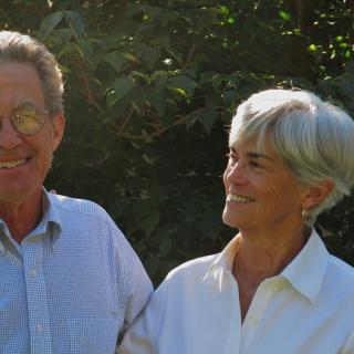 An elderly couple smiling at each other in a sunny outdoor setting.