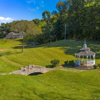 A scenic view featuring a gazebo, stone bridge, and a house set against a backdrop of lush greenery.