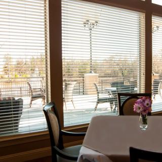 A cozy restaurant interior with a view of outdoor seating through large windows with blinds.