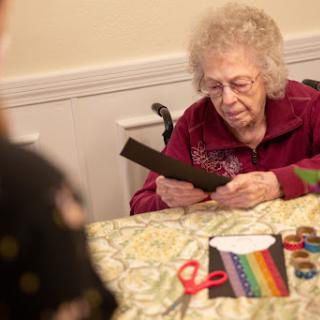 An elderly woman engaged in a crafting activity, focusing on colorful paper materials and scissors at a table.