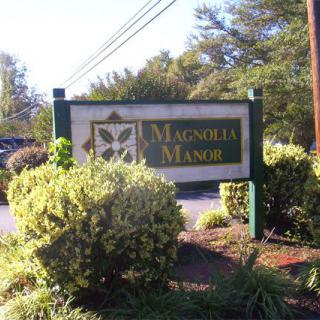 A sign for Magnolia Manor surrounded by greenery and bushes.