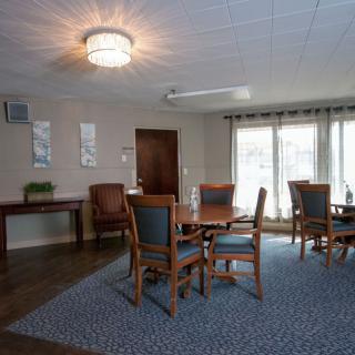A cozy dining area with wooden tables and chairs, illuminated by a ceiling light and natural light from large windows.