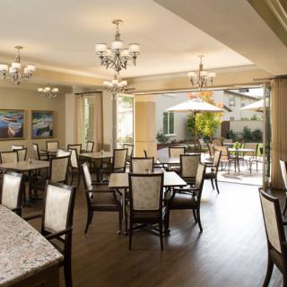 An elegant dining area featuring multiple tables and chairs, illuminated by chandeliers and open to a bright outdoor space.