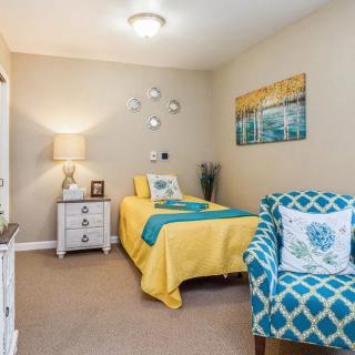 A cozy bedroom featuring a single bed with yellow and blue bedding, a patterned armchair, and decorative artwork on the wall.