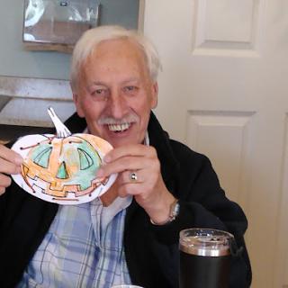 An elderly man proudly displaying his colorful pumpkin craft at a cozy indoor setting.