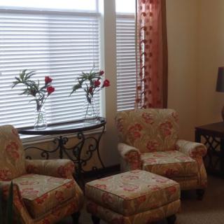 Cozy living room with floral-patterned chairs, matching ottomans, and a decorative table adorned with vases of flowers by the window.